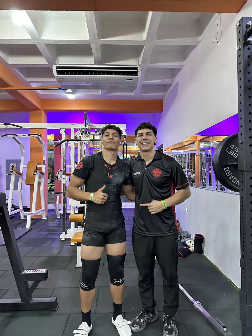 Dos atletas de Overlimit listos en la sala de pesas de la sede, con rodilleras y cinturones, antes de salir a la plataforma.